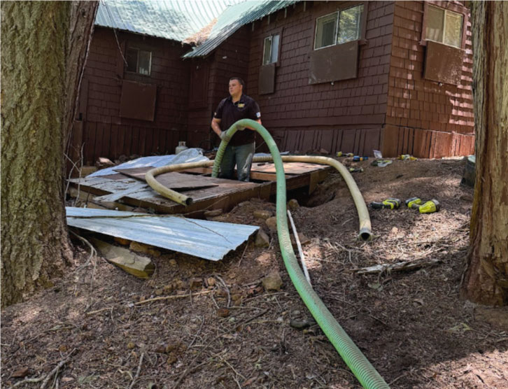 How Much Does Septic Tank Pumping Cost? Rescue Hero Plumbing technician pumping a septic tank buried beneath wooden planks in a wooded backyard, demonstrating difficult access for septic pumping cost factors.