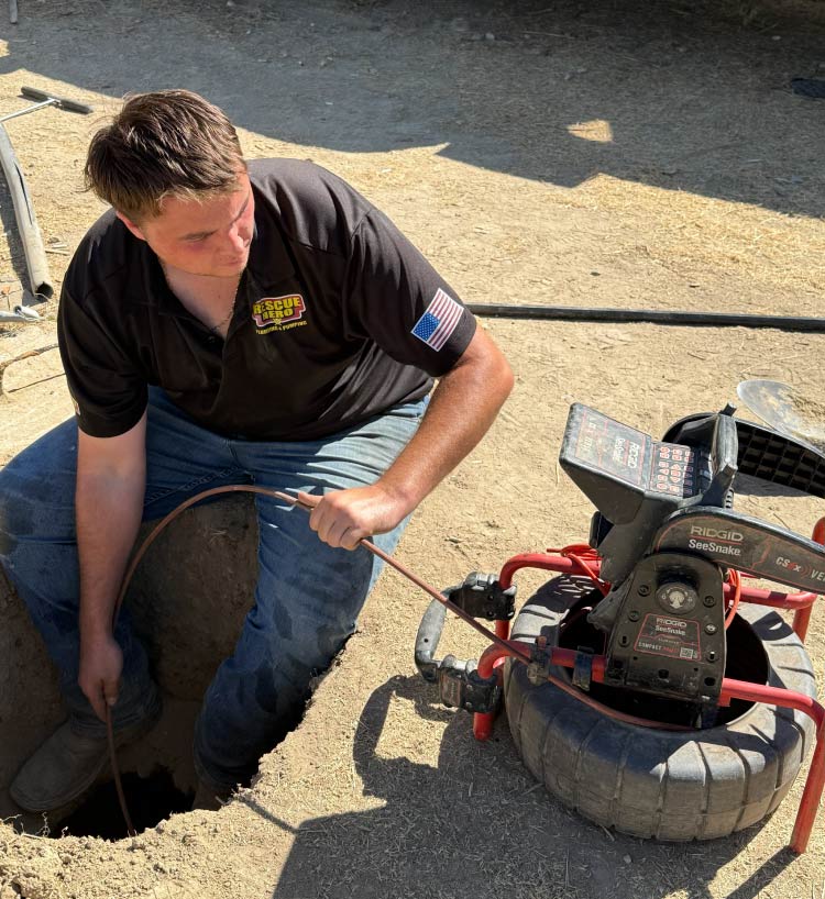 Plumbing technician utilizing a Ridgid SeeSnake video camera system to inspect an underground sewer line for blockages, roots, or leaks within a dirt trench.