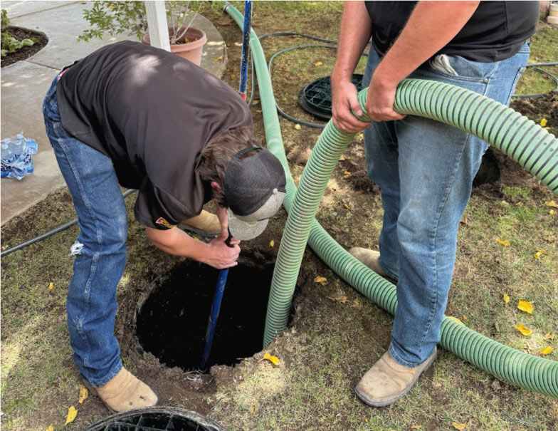 Two Rescue Hero Plumbing technicians working together to carefully guide a heavy-duty green vacuum hose into an open underground septic tank for routine pumping and cleaning in a residential yard.