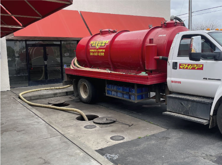 Rescue Hero Plumbing red pumping truck hooked up to commercial-sized septic tanks with manhole covers in a paved lot, showing commercial septic pumping service.