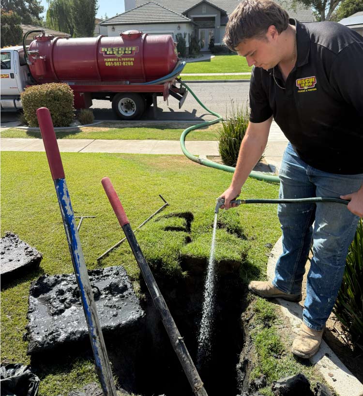 Technician using a hose to clean a freshly excavated septic tank access point in a residential front yard, with a Rescue Hero Plumbing pumping truck visible in the background