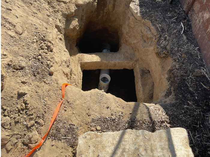 Deep excavation exposing the inlet and outlet PVC pipes of a concrete septic tank for necessary repairs.