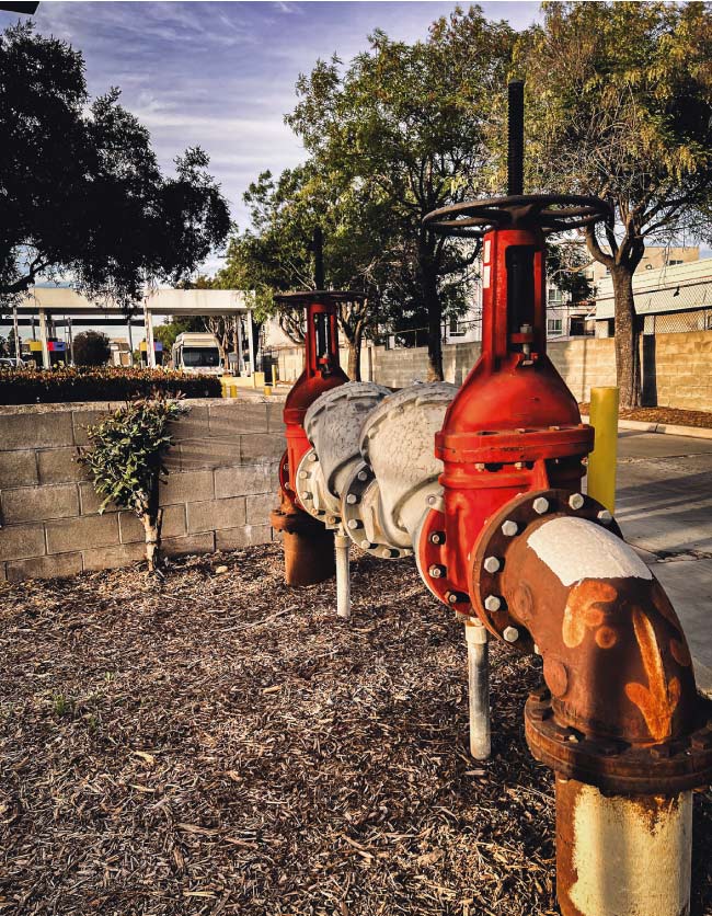 Large red reduced pressure zone (RPZ) backflow prevention assembly with two shut-off valves installed near a commercial parking lot wall to prevent cross-contamination.