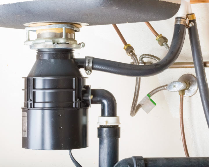 Close-up of a black garbage disposal unit installed under a kitchen sink with copper water lines and black rubber discharge hoses, often inspected during professional garbage disposal repair.