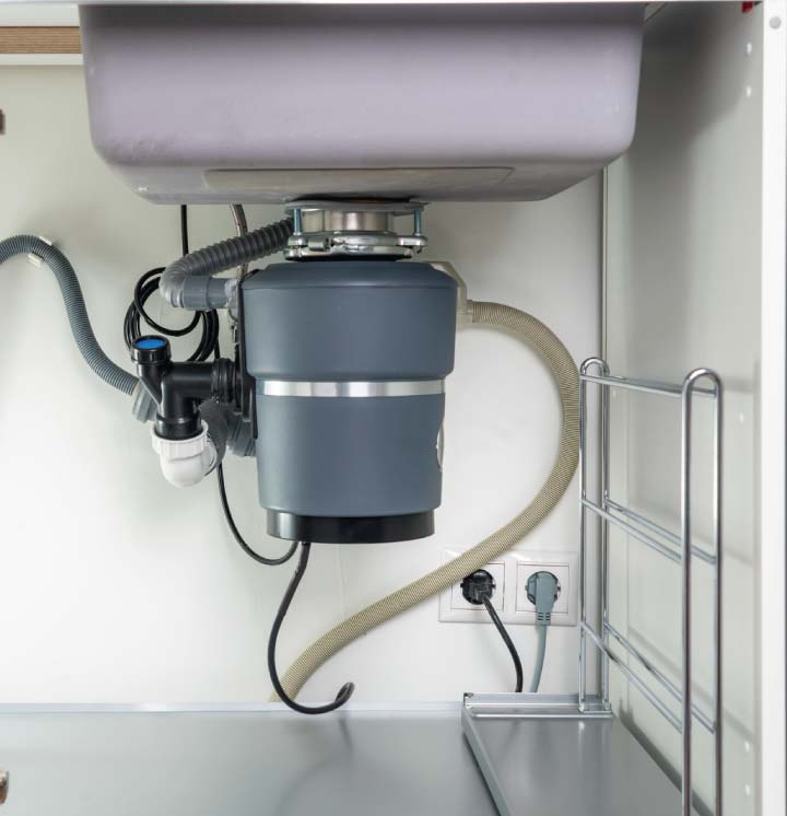 Modern grey garbage disposal mounted under a stainless steel sink with a white P-trap and dishwasher drain connection, demonstrating a clean and proper installation.
