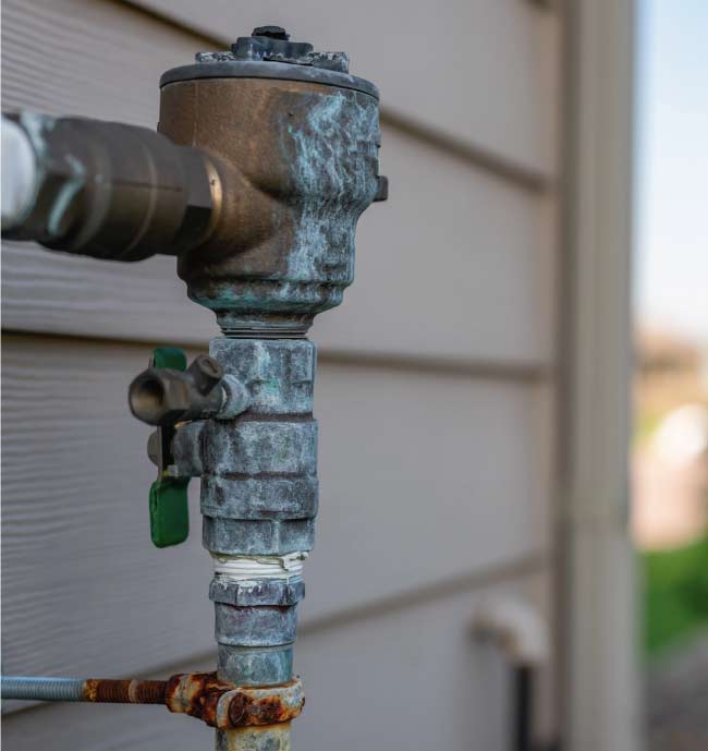 Residential pressure vacuum breaker (PVB) backflow device attached to the side of a home with tan siding, showing signs of oxidation and ready for a professional inspection.