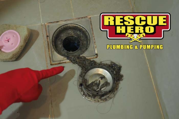 Red gloved hand pointing to a massive clump of black hair and sludge pulled from a square stainless steel shower drain cover during a bathroom cleaning.
