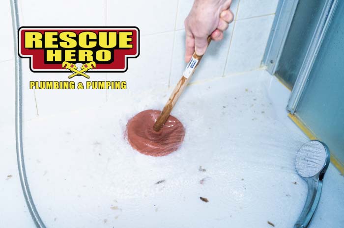 Person holding a wooden-handled cup plunger over a soapy, water-filled shower floor to clear a blockage, featuring the Rescue Hero Plumbing logo.