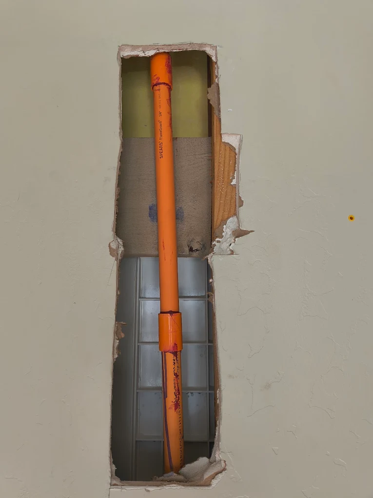 Vertical orange water pipe with a coupling exposed inside a cut-out section of drywall, illustrating a plumbing line repair or repiping project behind a finished wall.
