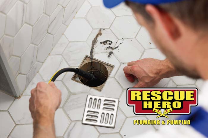How to Fix a Clogged Shower Drain: Professional plumber feeding a black mechanical snake cable into a square floor drain surrounded by white hexagonal tiles to clear a deep pipe blockage.