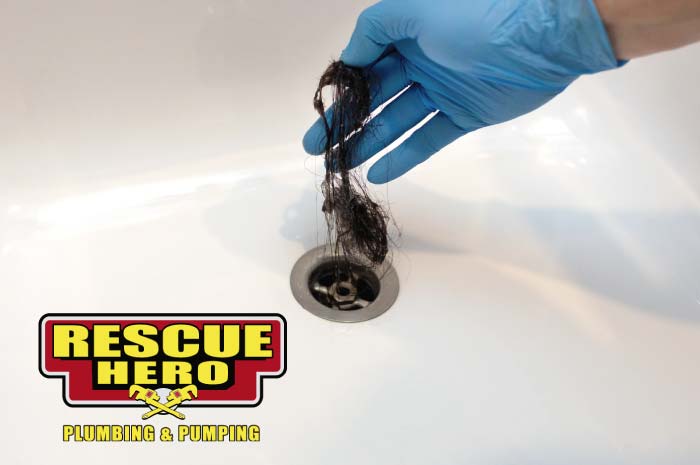 Person wearing a blue nitrile glove holding a long clump of dark hair retrieved from a white shower drain, illustrating the most common cause of bathroom clogs.