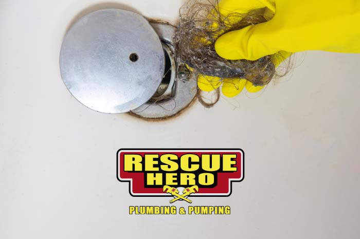 Close-up of a hand in a yellow rubber glove removing a large tangle of hair and soap scum from a circular chrome shower drain to fix a slow drain issue.