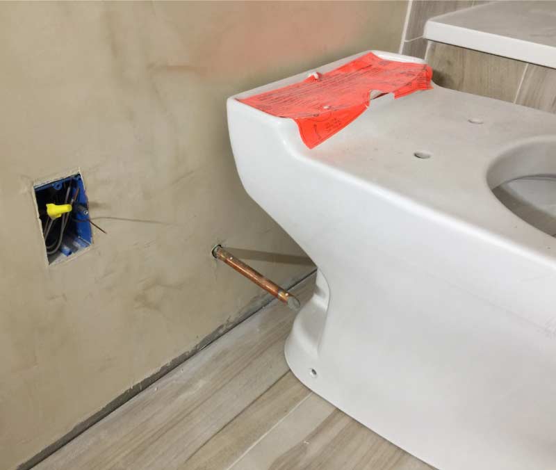 New white toilet unit being installed on wood-look tile flooring, positioned next to a copper water supply line and an electrical box for a potential bidet attachment.