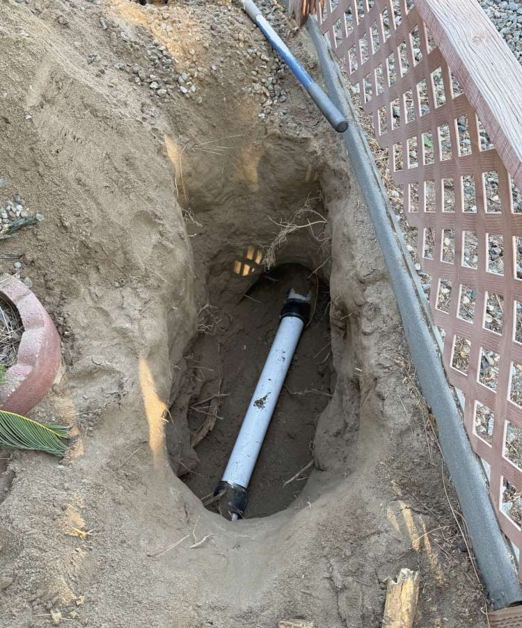 Newly installed section of white PVC sewer pipe secured with black flexible couplings, exposed inside a deep dirt trench during an underground residential plumbing repair next to a wooden fence.