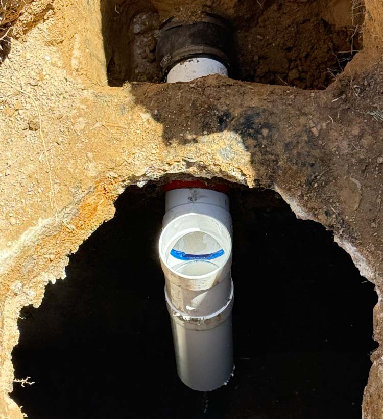 A top-down view of a Whole-House Repiping project in progress, showing new white PVC plumbing pipes connected with a red coupler and black rubber gasket inside a excavated dirt trench