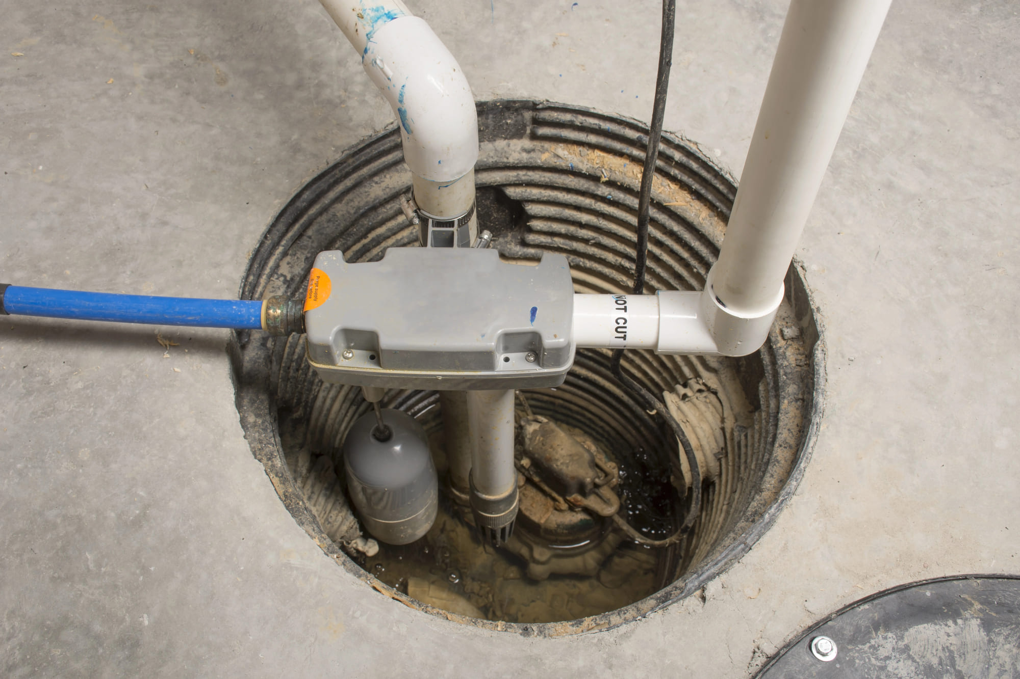 A detailed view of a modern residential sump pump system installed in a circular basin within a concrete floor, featuring a gray pump housing, a blue water line, white PVC piping, and a visible gray float switch.