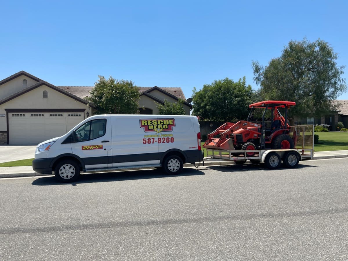 White Rescue Hero Plumbing and Pumping service van parked on a residential street, towing a flatbed trailer with an orange mini-excavator used for underground sewer and septic tank installations.