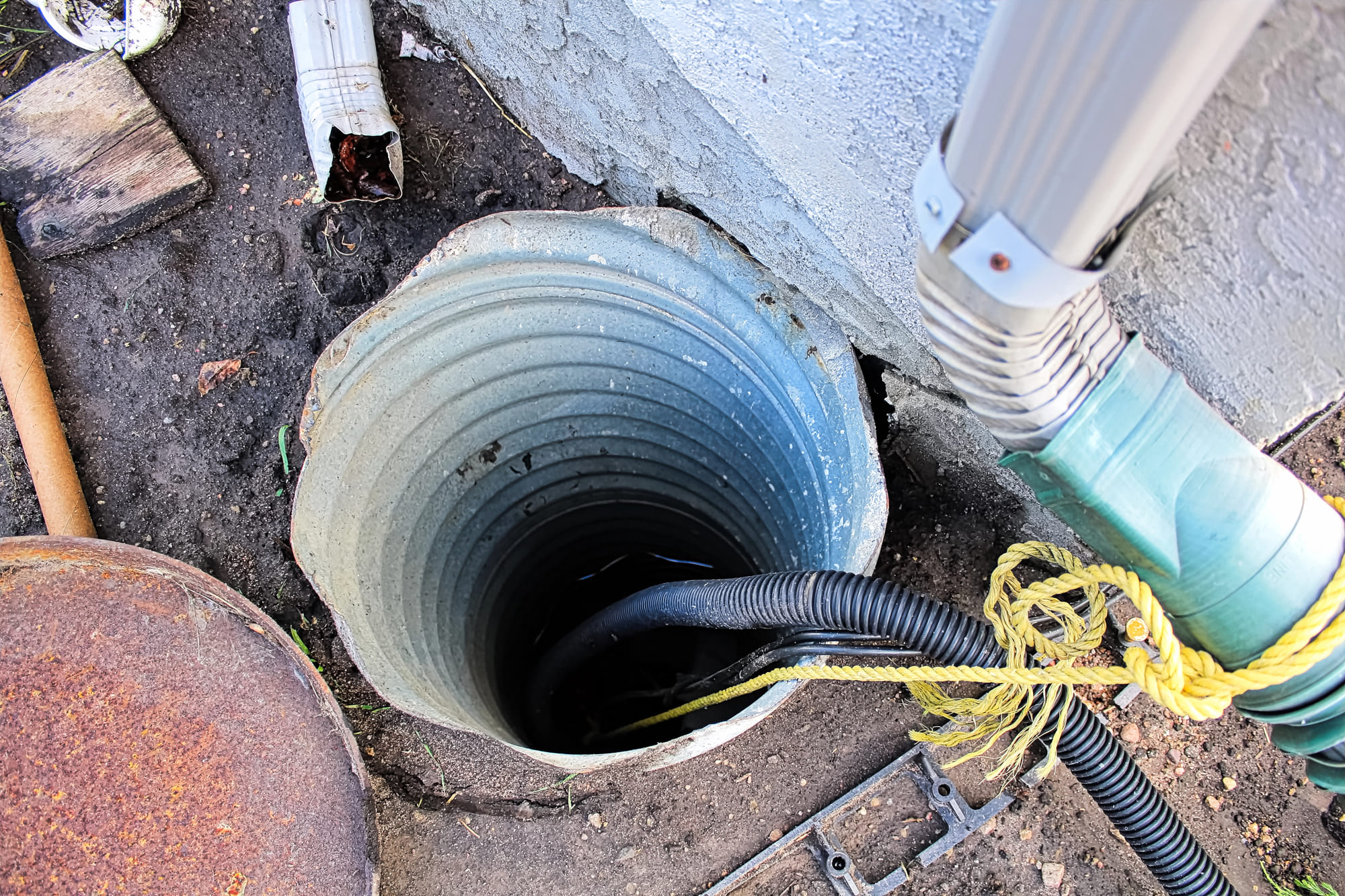 An outdoor view of a metal sump pump basin or well casing recessed into the ground next to a building foundation, with a black corrugated discharge hose and a yellow pull rope visible inside the pit for sump pump services