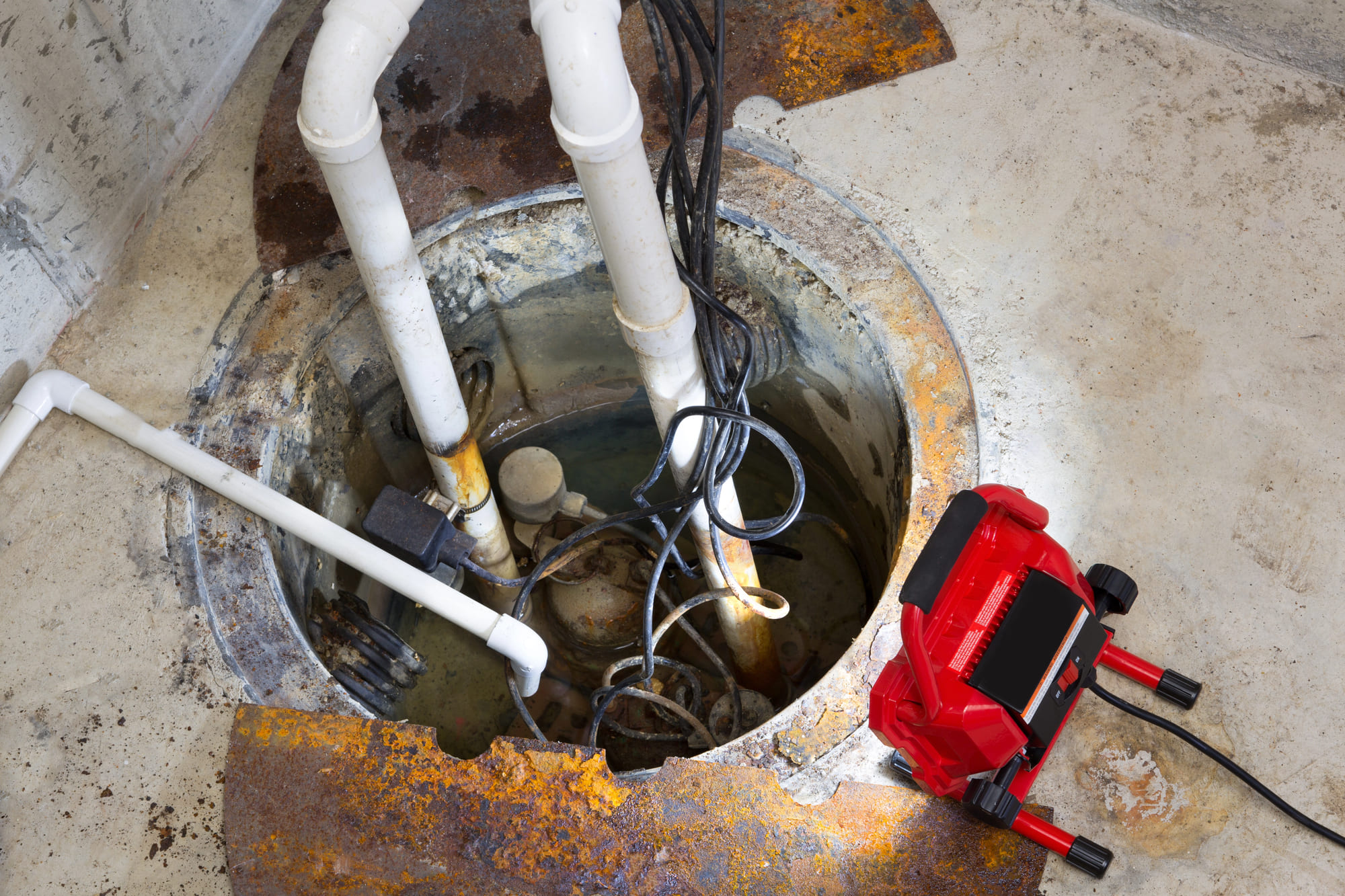 A high-angle, close-up shot of a dirty residential sump pump pit in a concrete floor, showing the pump, white PVC discharge pipes, and tangled black power cables being illuminated by a bright red portable LED work light.